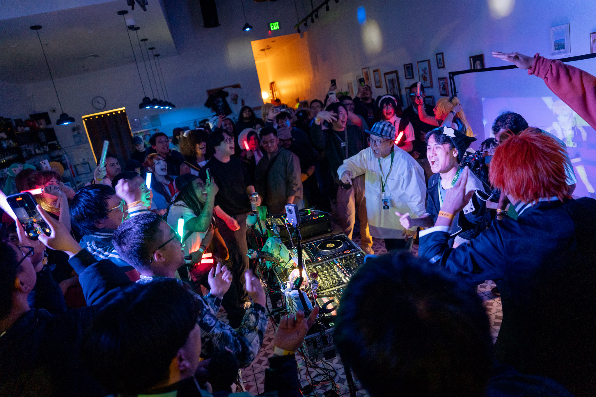 A room of people surrounding a dj, some holding pen lights, plushes, everyone smiling and hyped